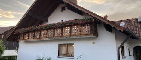 Ein traditionelles Chalet mit einem geneigten Dach, einem hölzernen Balkon mit Blumenkästen und einem kleinen Fenster.