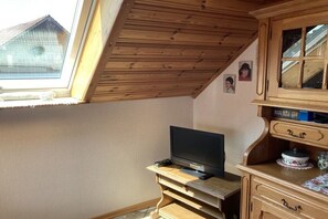 Een kamer met een schuine kap, een televisie op een houten standaard, een houten kast met lades en een raam met uitzicht op een huis.