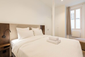A hotel room with a large bed, a bedside table, a wall-mounted lamp, and a window with curtains. - Elégant appartement près du Moulin Rouge (Paris)