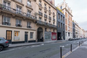 A row of multi-story buildings with balconies, a street with parked cars, and a pedestrian walkway. - Elégant appartement près du Moulin Rouge (Paris)