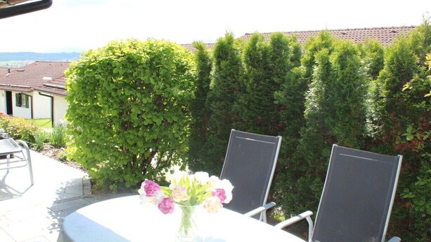 Eine Terrasss mit Tisch und zwei Stühlen, einem Blumenstrauß und einem Hecken im Hintergrund.