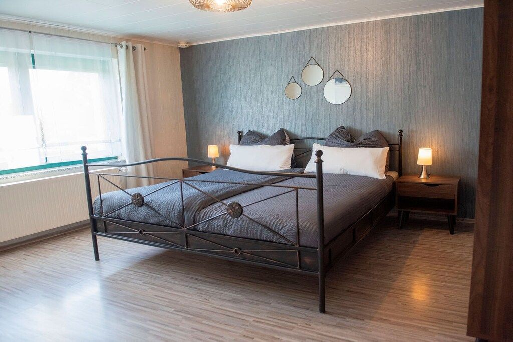 A bedroom with a metal bed frame, two bedside tables with lamps, a window with curtains, and a wall-mounted mirror.