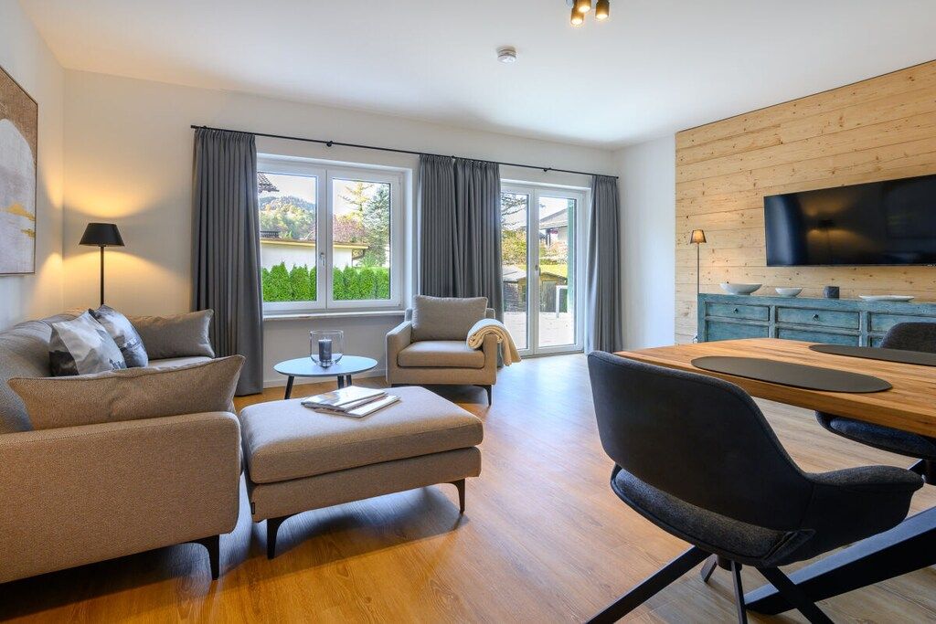 Ein modernes Wohnzimmer mit einer Couch, einem Sessel, einem Hocker und einem Esstisch mit Stühlen. Ein Fernseher ist an der Wand montiert und ein Fenster mit Vorhängen vorhanden.