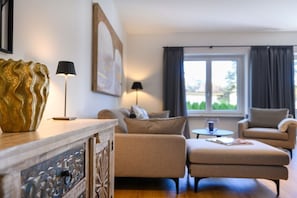 A modern living room with a sofa, armchair, and ottoman, a decorative gold vase, and a lamp on a wooden console.