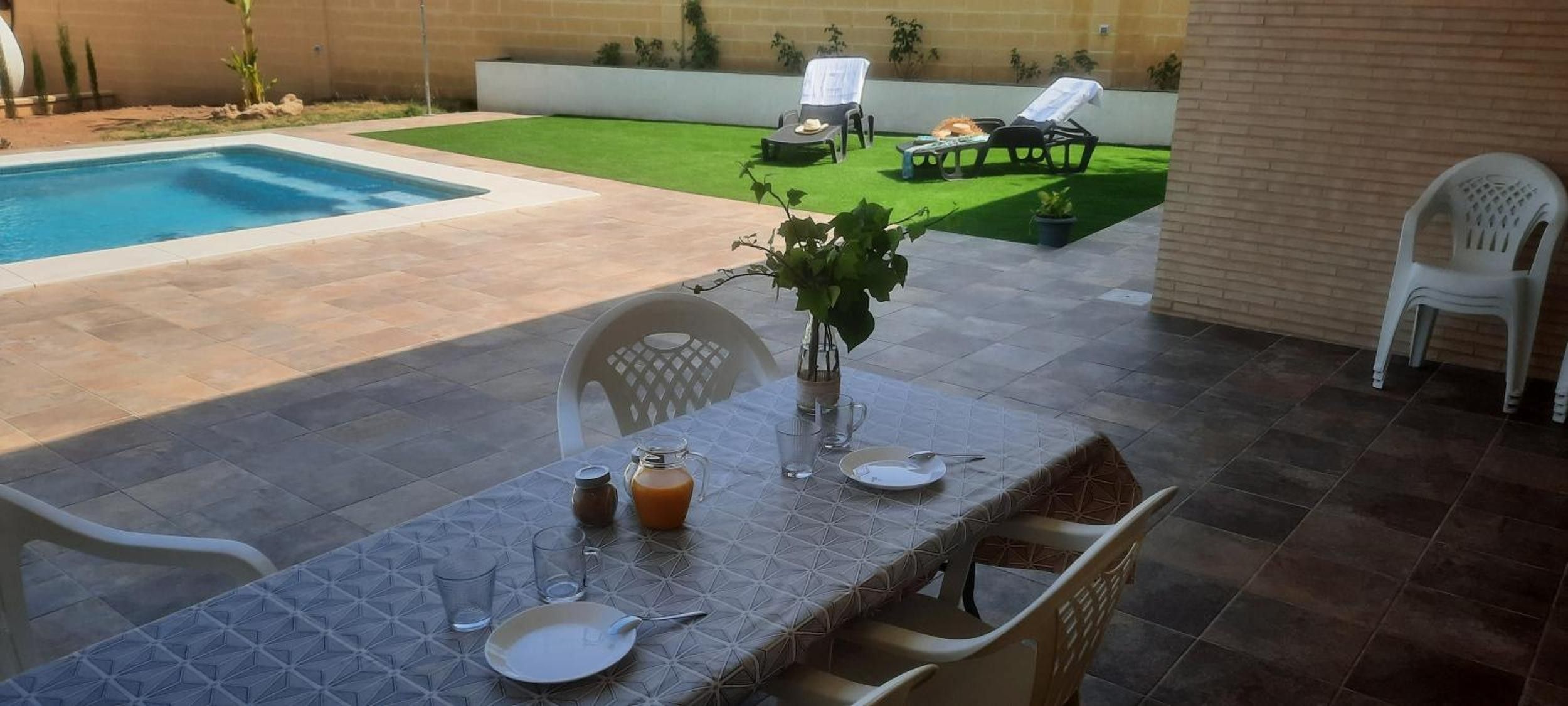 Comedor con mesa lista para dos, piscina y jardín con sillones de descanso.