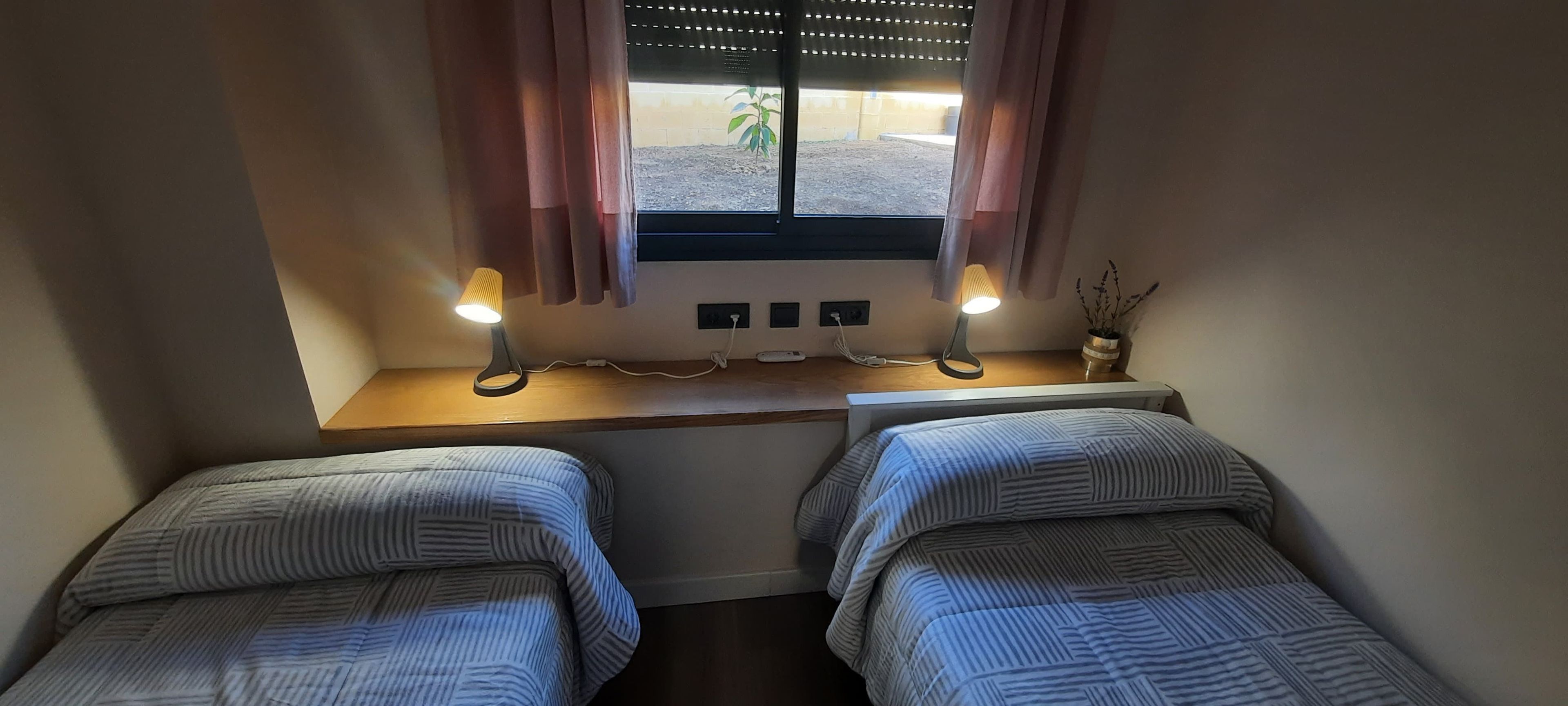 Ein kleines Zimmer mit zwei Betten, einem Fenster mit Vorhängen und zwei Nachttischlampen.