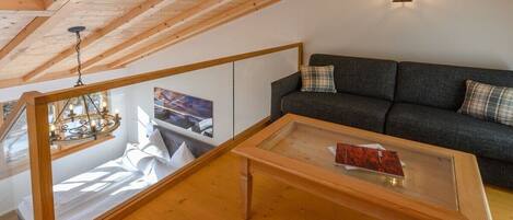 A loft-style room with a wooden ceiling, a glass table, a sofa, and a hanging light fixture.