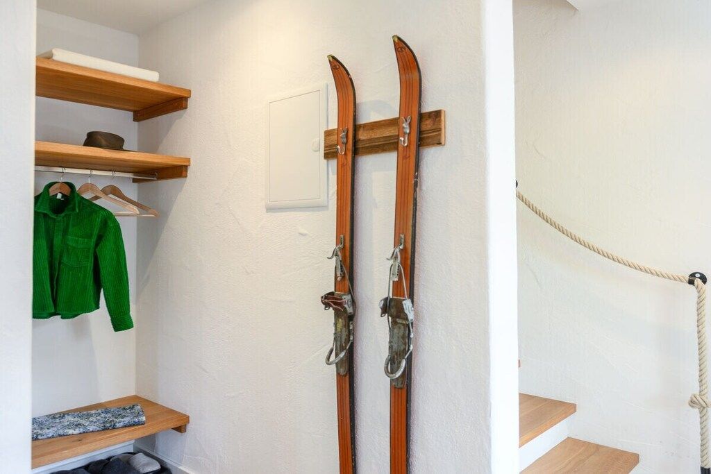 A room with a wooden wall-mounted ski rack, a coat rack with a green jacket, and a rope tied to the wall.