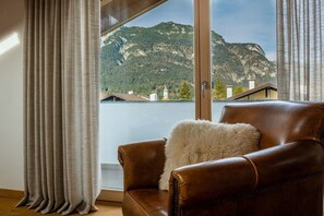 Ein Zimmer mit Ledersessel, einem flauschigen Kissen und Blick auf Berge und See durch ein Fenster.