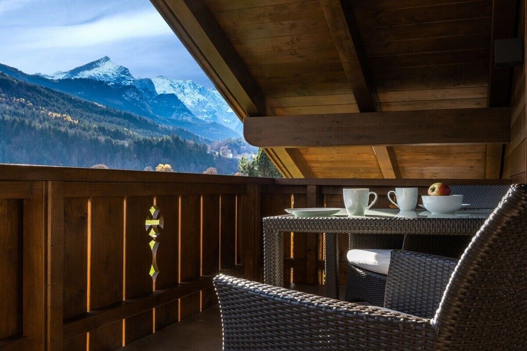 A wooden balcony with wicker furniture, a table with cups and a bowl, and a mountain view.
