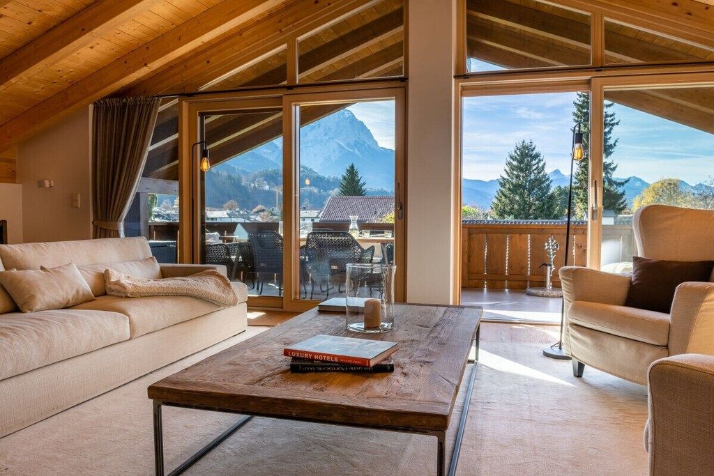 A spacious living room with a wooden ceiling, large windows offering mountain views, a sofa, a coffee table, and a chair.