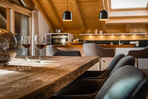 Ein Holztisch mit zwei Gläsern Rotwein, einer strukturierten Schale und modernen Stühlen in einer Küche mit Holzbalken und Pendelleuchten.