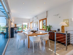 Ein geräumiger Essbereich mit einem Holztisch und weißen Stühlen, eine Küche mit Marmorarbeitsplatte und ein Blick auf eine grüne Landschaft durch große Fenster.
