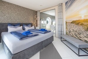 Ein modernes Schlafzimmer mit einem großen Bett, einer Bank, einem Badezimmer mit Spiegel und einer Wand im Strandhausstil.