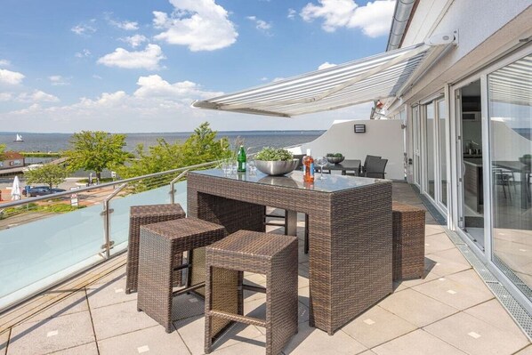 Ein Balkon mit Korbmöbeln, einem Glastisch und Blick aufs Meer.