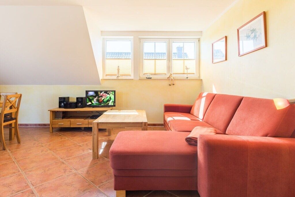 Un salon avec un canapé rouge, une table basse en bois, un téléviseur posé sur un meuble et une fenêtre avec des stores.