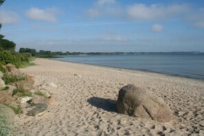 Een zandstrand met een grote rots op de voorgrond en een rustig waterlichaam op de achtergrond.