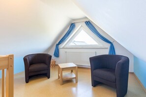 A room with a sloped ceiling, a window with blue curtains, two dark-colored chairs, and a small wooden table. - B - Holiday home Katharina (Wohlenberg)