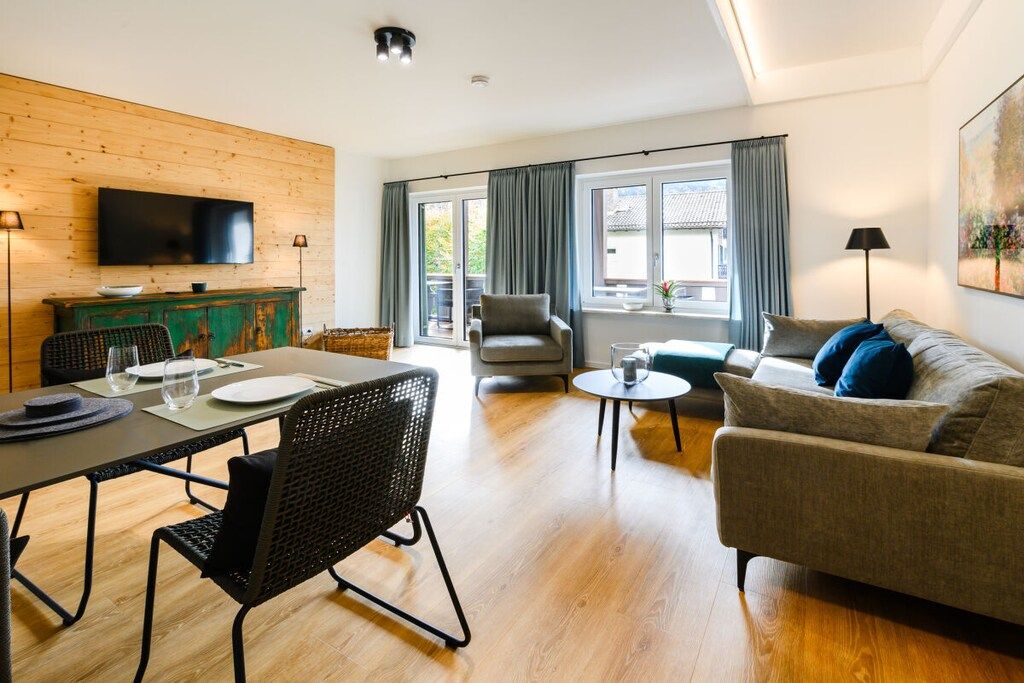 Ein modernes Wohnzimmer mit Essbereich, einem an einer Holzwand montierten Flachbildfernseher, einer Couch, einem Sessel und einem kleinen runden Tisch.