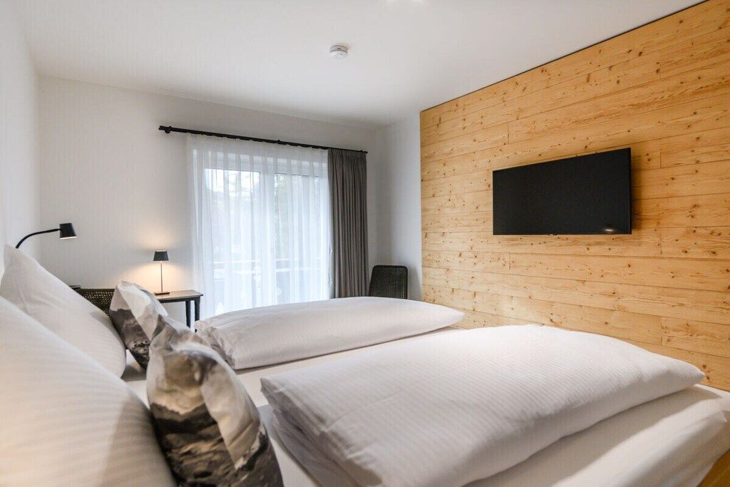 Une chambre d’hôtel avec un mur en bois, un téléviseur à écran plat, un lit avec du linge de maison blanc, un bureau avec une lampe et une fenêtre avec des rideaux légers.