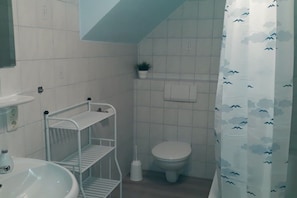 Kylpyhuoneessa on valkoinen pesuallas, wc-istuin, metallinen hyllykkö, pilvikuvioitu suihkuverho ja pieni ruukkukasvi seinällä.