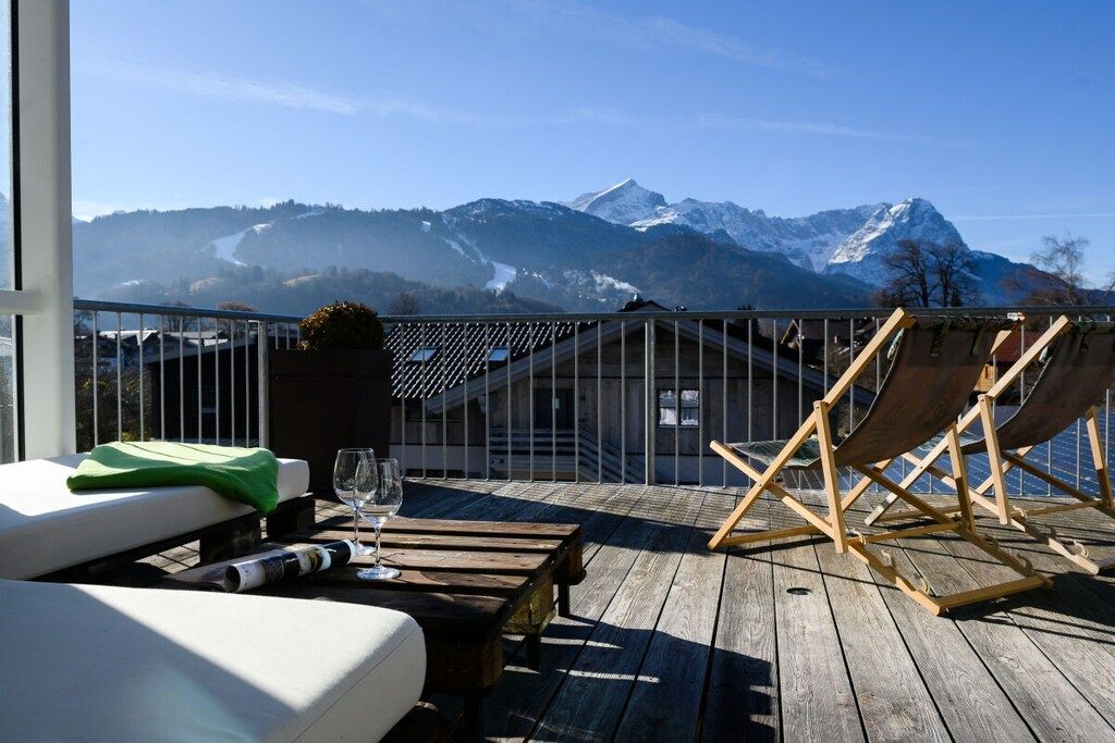 A wooden deck with a lounge chair, a table with a glass, and a view of snow-capped mountains.