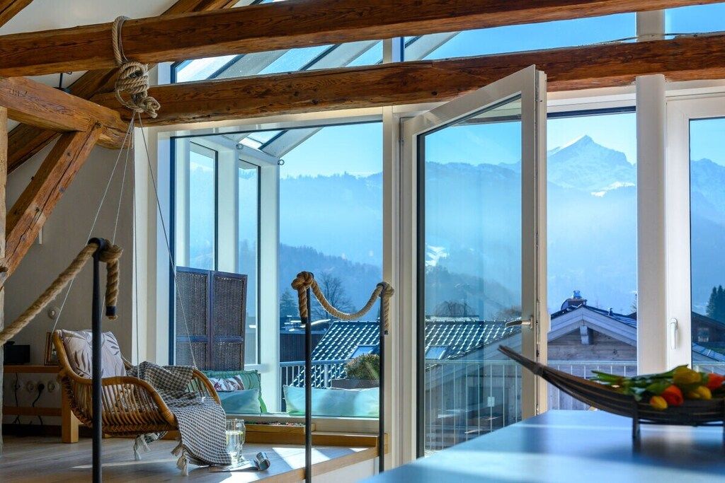 A room with a large window, wooden beams, a hanging chair, and a view of mountains.