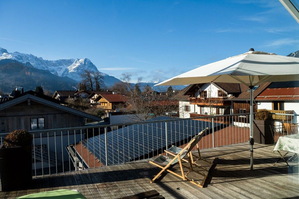 A rooftop terrace with solar panels, a lounge chair, and a table under a large umbrella, overlooking a mountain range.