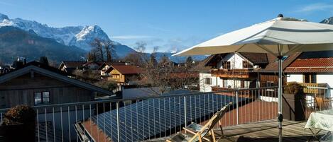 A rooftop terrace with solar panels, a lounge chair, and a table under a large umbrella, overlooking a mountain range.