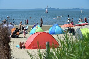 Een strand met tenten en parasols, mensen zwemmen en wandelen op het zand, en zeilboten op het water.