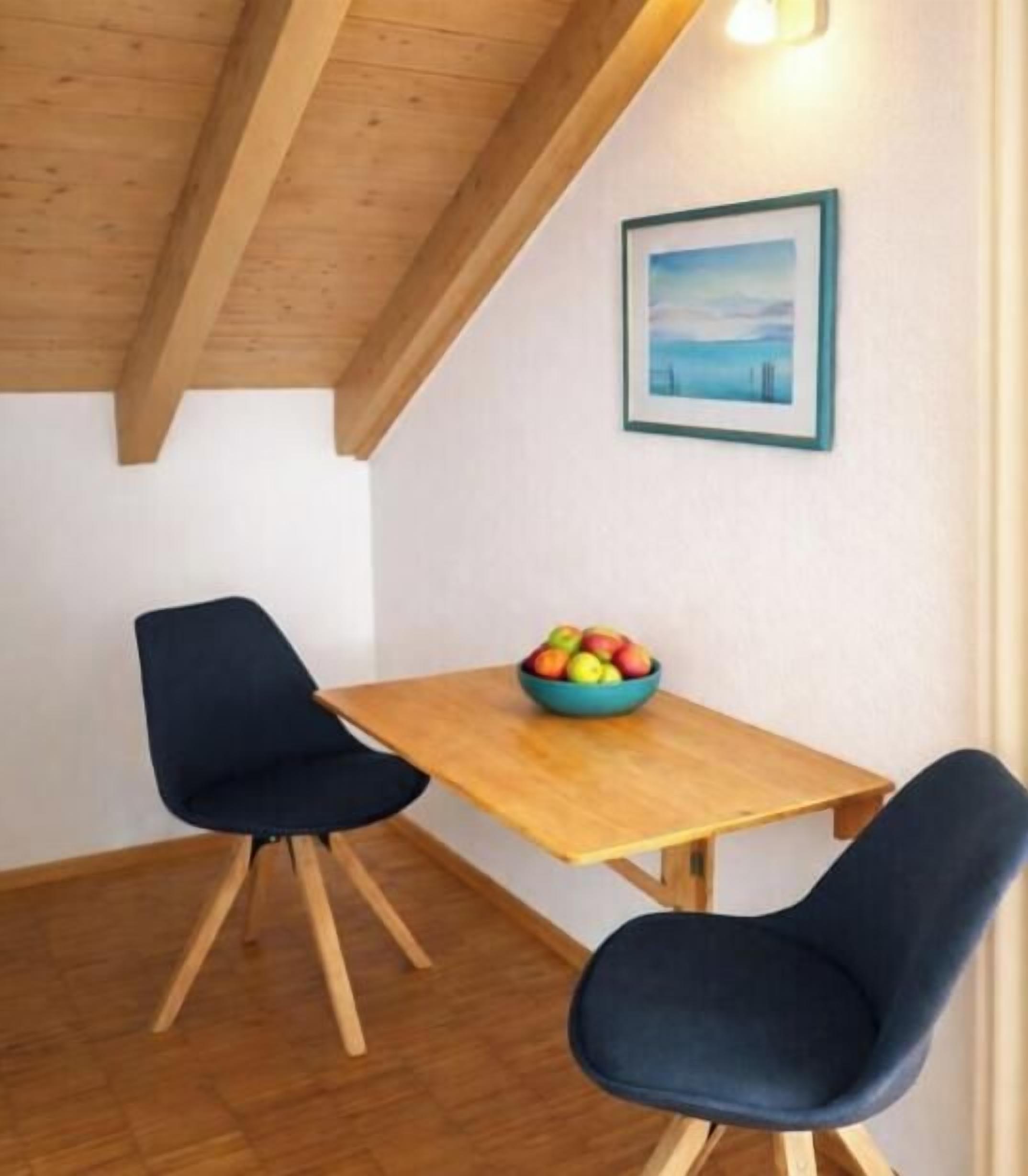 A dining area with a wooden table and two chairs, a bowl of fruit on the table, and a framed picture on the wall.