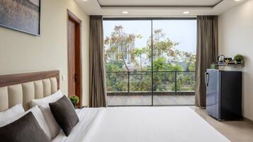 A hotel room with a large bed, a view of the city through a balcony, and a refrigerator.