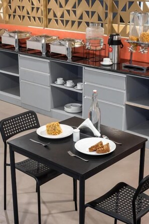 Uma sala de jantar com mesa posta para dois, um buffet com diversas opções de comida e um dispensador de água.