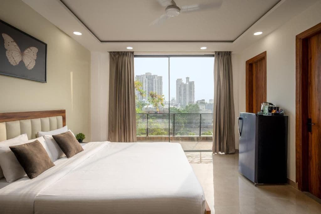 A hotel room with a large bed, a ceiling fan, a view of the city through the window, and a refrigerator.