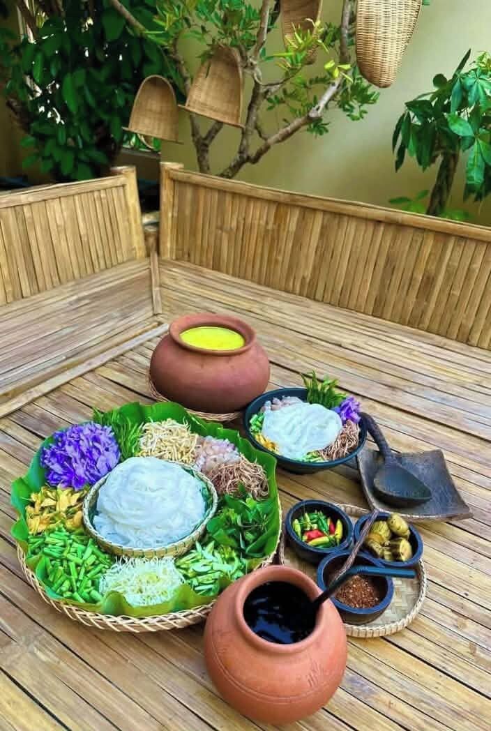 A traditional meal setup with various dishes and utensils on a wooden table, surrounded by greenery.