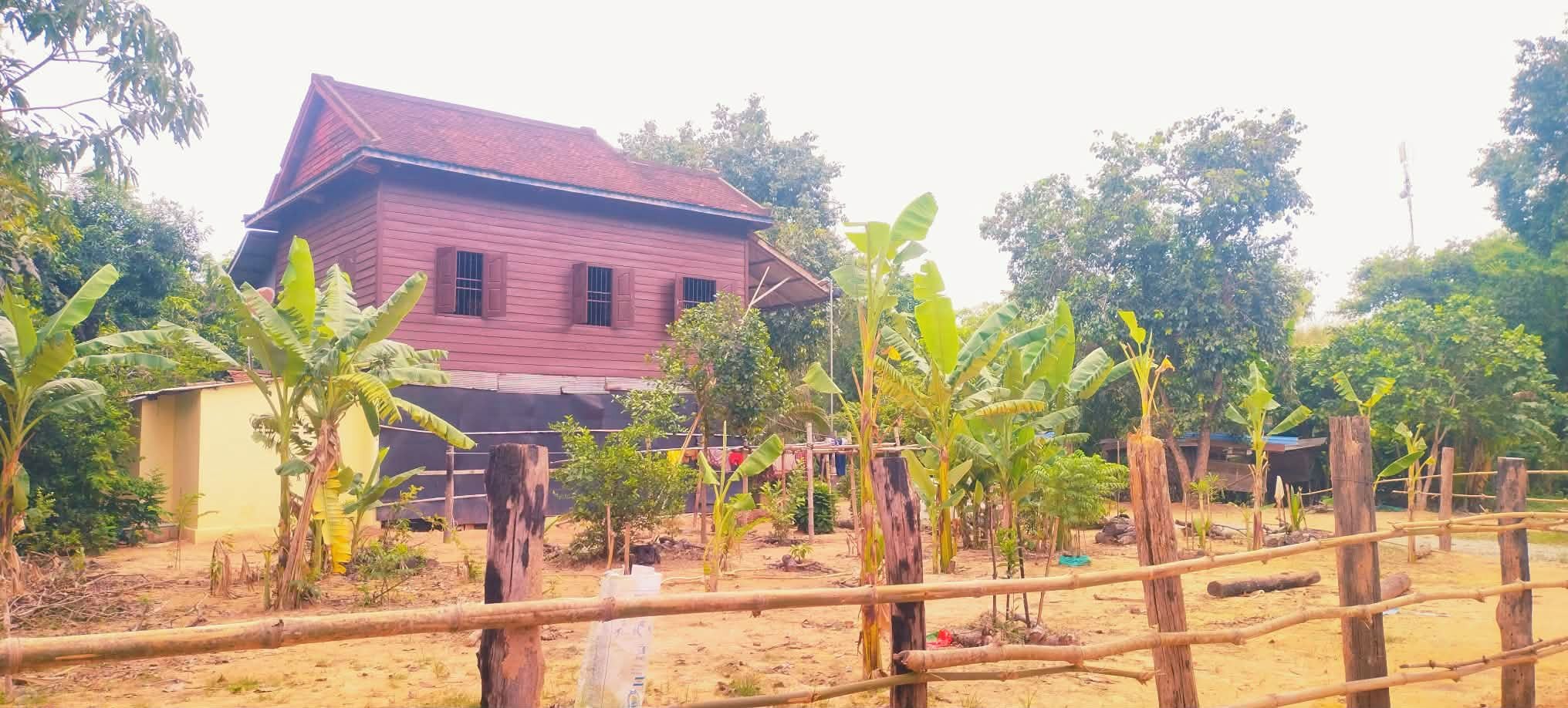 Uma casa vermelha com telhado inclinado, cercada por bananeiras e uma cerca de madeira.