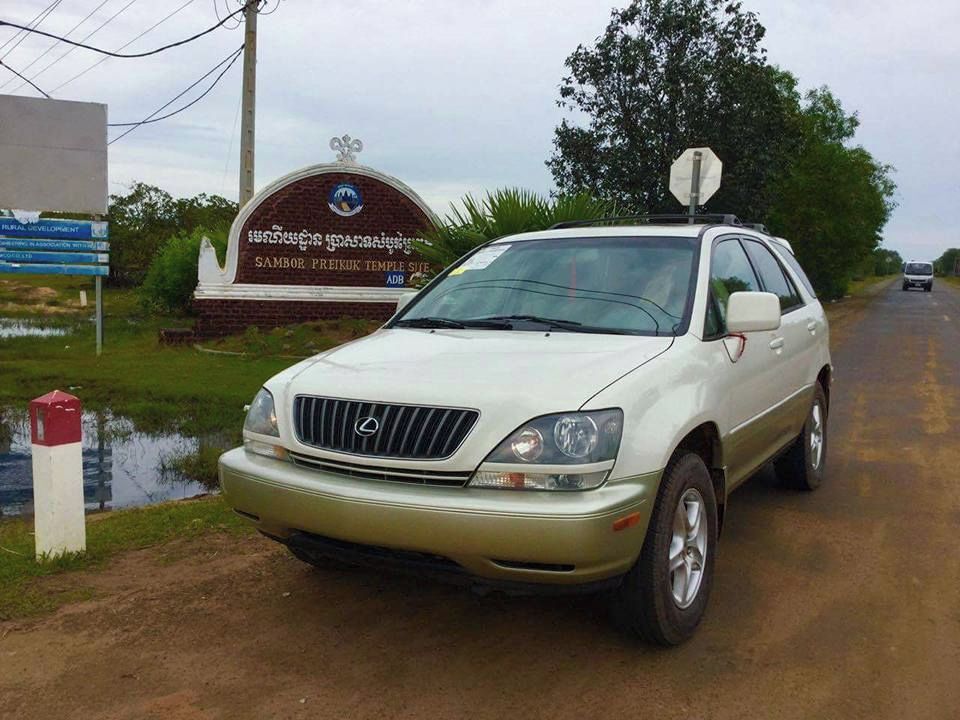 Um SUV branco estacionado em uma estrada rural, com uma placa indicando 'Sambor Preuk Tempel Sute' ao fundo.