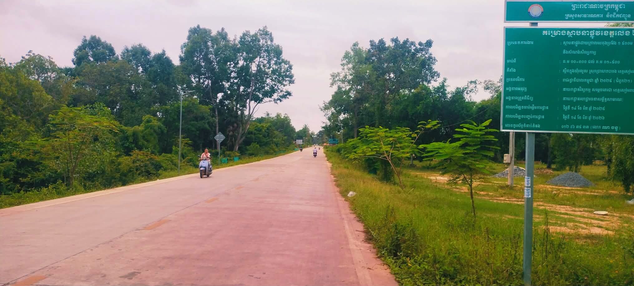 A paved road with trees on both sides, a signboard, and a motorcyclist in the distance.
