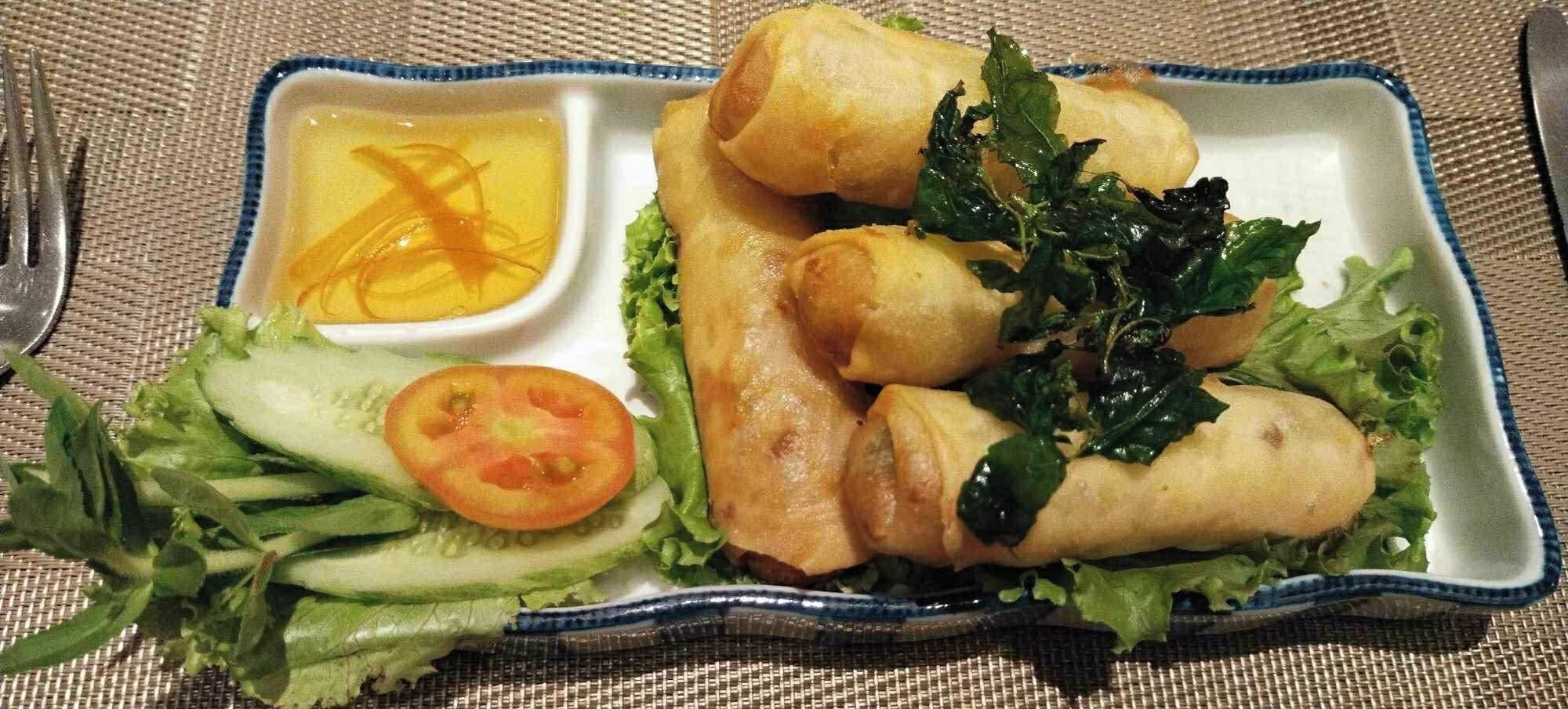A plate with spring rolls, lettuce, a slice of tomato, and a small bowl of sauce.