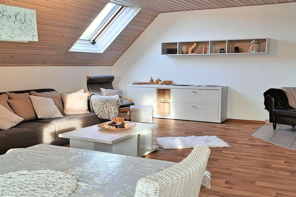 A modern loft-style living room with a sloped ceiling, a sofa set, a coffee table, a white cabinet, and a small shelf with decorative items.