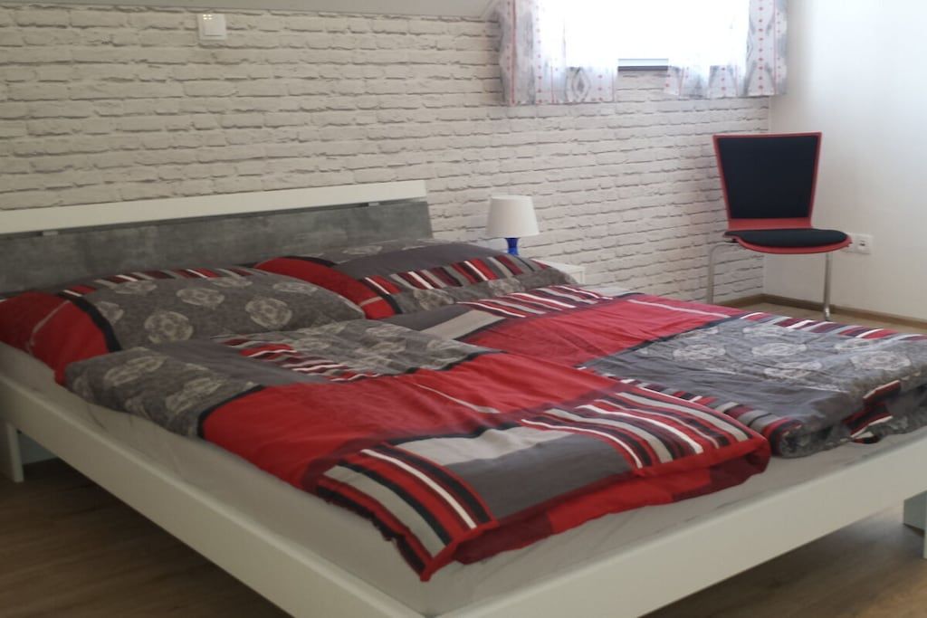 A bed with a white frame, a grey and red patterned bedspread, and a red and grey pillow. There is a white lamp on a bedside table and a red and black chair.