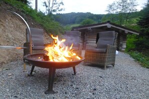 Un foc a terra amb flames, envoltat de mobles de vimet i un edifici petit en un entorn natural.