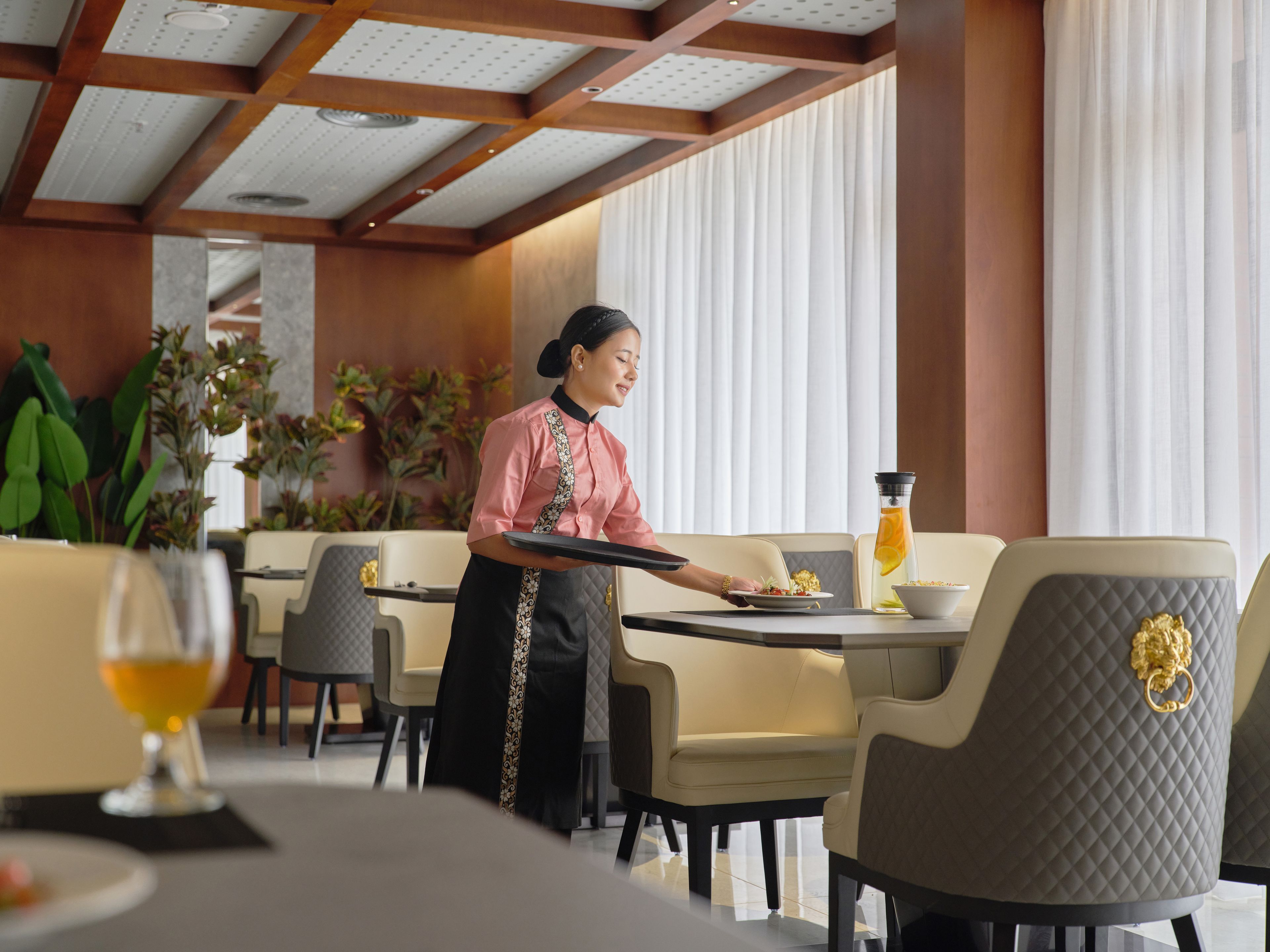 A hotel dining area with a waiter serving a guest. There are cushioned chairs, a table with a glass top, and a bottle of orange juice.