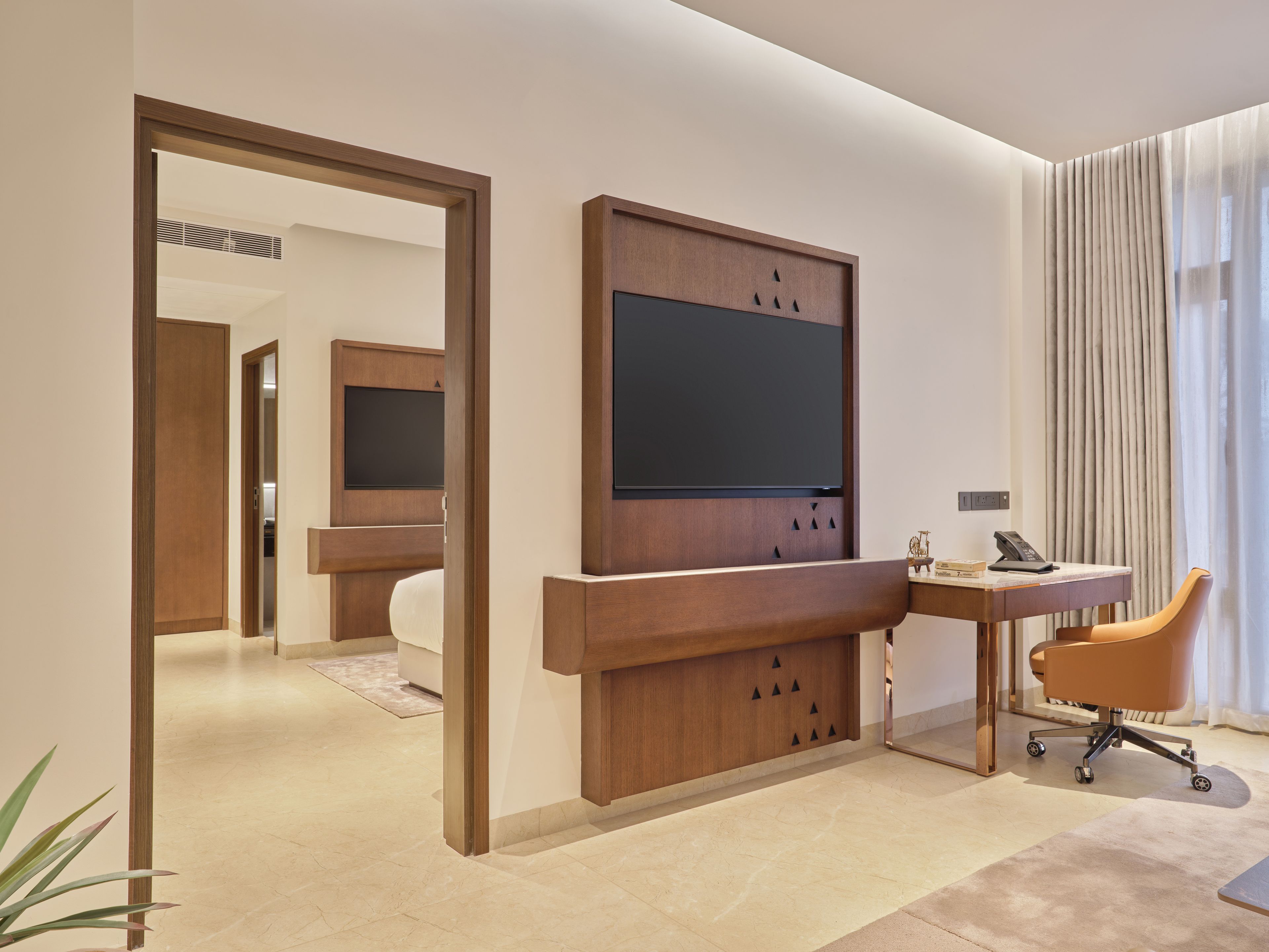 A modern hotel room with a large flat-screen TV mounted on a wooden panel, a desk with a telephone, and a comfortable chair.