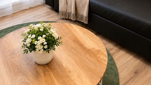 Uma sala de estar moderna com um sofá de couro preto, uma mesa de centro redonda de madeira com uma planta em vaso e um tapete verde.