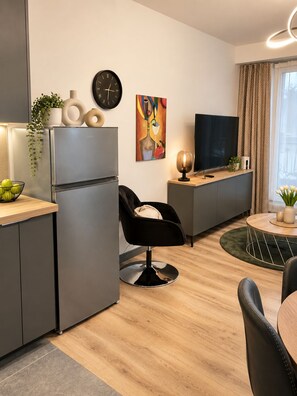 Una cocina moderna con refrigerador incorporado, un comedor con mesa redonda y sillas, una silla negra y un televisor de pantalla plana sobre un pedestal de madera.