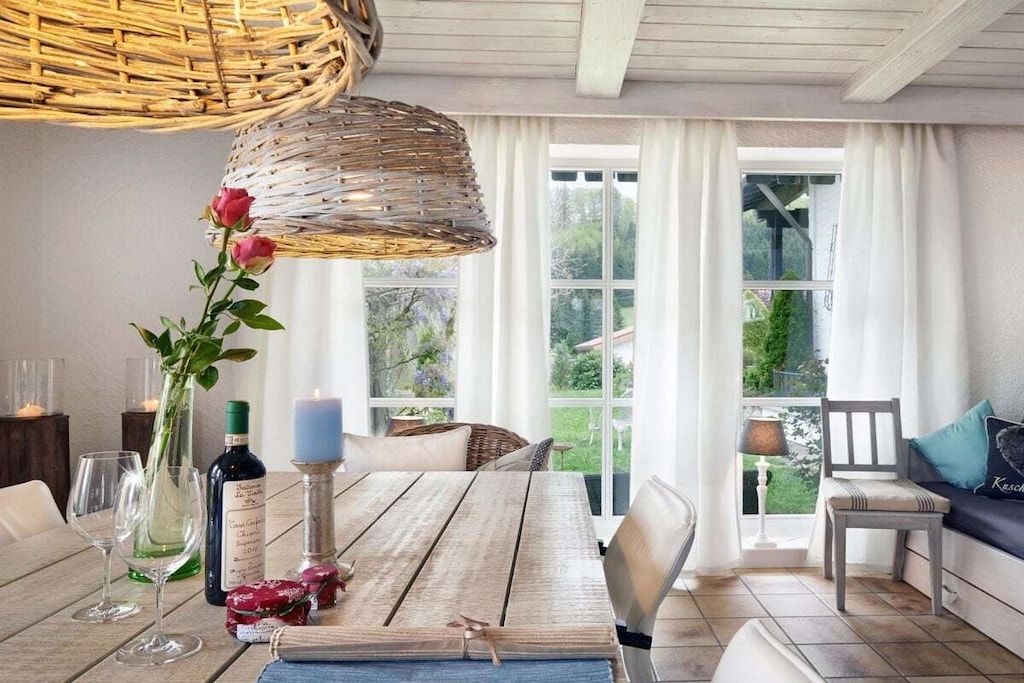Un comedor con mesa de madera, sillas, lámpara colgante de mimbre, un florero con flores y vistas al exterior.
