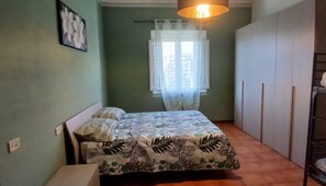 Un dormitorio con cama, una ventana con cortinas translúcidas, un armario y una lámpara fijada a la pared.