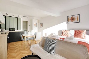 Quarto de hotel moderno com sofá, cama com almofadas decorativas, área de jantar com mesa e cadeiras, e cozinha compacta com micro-ondas.