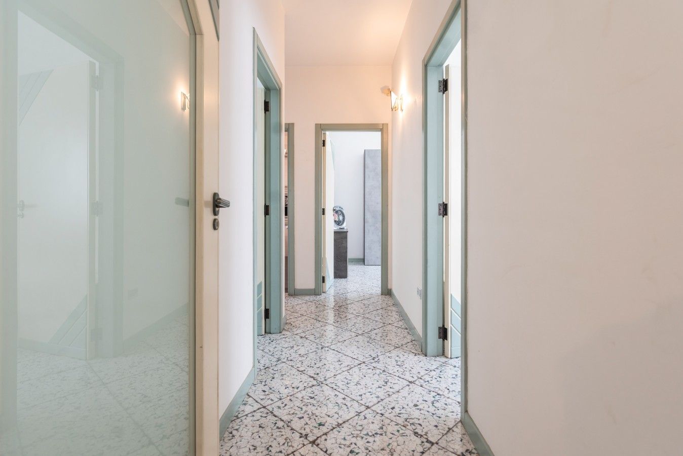 A corridor with a tiled floor, doors on both sides, and a light fixture on the wall.